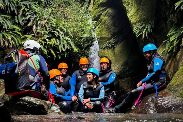 Group in helmets and wetsuits sitting by waterfall amid lush greenery.