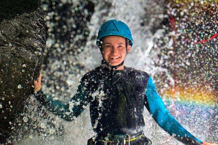 Person in wetsuit and helmet standing in waterfall spray, smiling.