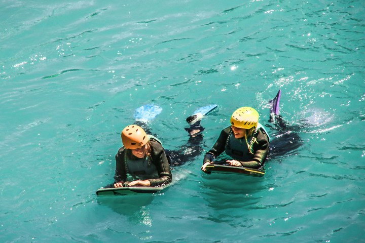 Two people with helmets floating on turquoise water using bodyboards.