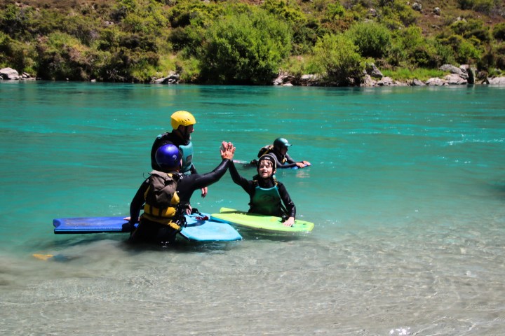 People in helmets and wetsuits high-five in a river with bodyboards, surrounded by greenery.