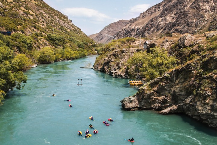 People rafting in a wide, turquoise river between rocky, green hills under a clear sky.