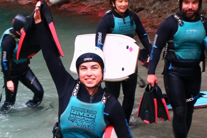 Group of four people river surfing in wetsuits and helmets, holding boards and flippers.