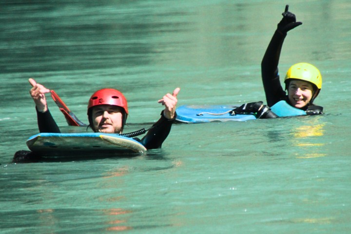 Two people in helmets swimming in a river with bodyboards, smiling and giving thumbs up.
