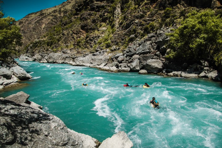 People riverboarding in a turquoise mountain river with rocky banks.