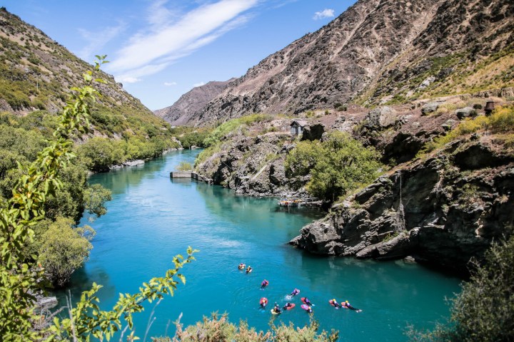 Swimmers in a blue river surrounded by rocky mountains and green vegetation.